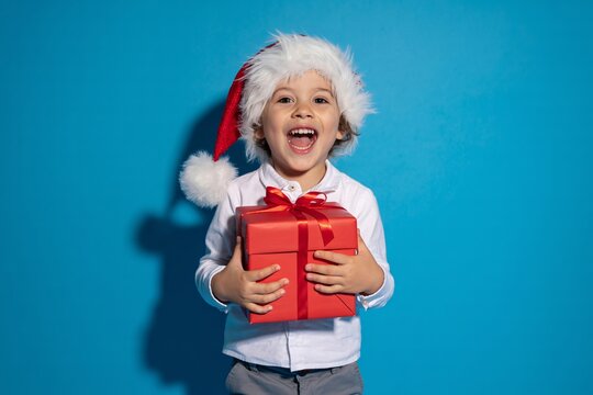 Photo of excited young boy wearing a santa hat, holding a red gift box with a bow, smiling widely against a vibrant blue background, embodying the joy of christmas