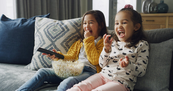Naklejki Happy girl, sisters and watching TV with popcorn on sofa for funny cartoon or movie in home. Child, siblings or kids laughing with snack or remote for online entertainment or subscription in house