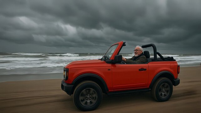 Smiling senior man driving a red convertible along a sandy beach under stormy clouds, enjoying freedom and coastal adventure.