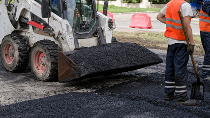 Skid steer loader unloads hot asphalt as road crew in safety vests spread mix on a city street...