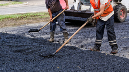 Unrecognizable road crew spreads and levels fresh asphalt by hand with rake and shovel during...
