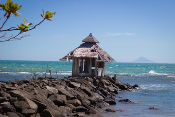 Scenic view of an abandoned wooden hut with thatched roof standing on a rocky shore, overlooking the turquoise sea under a clear blue sky. A distant island is visible on the horizon