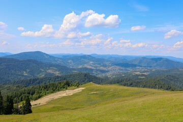 Naklejka premium A summer mountain landscape where the foreground is a green hill, offering a breathtaking panorama of wooded mountain ranges. In the distance, surrounded by greenery, a small village is visible.