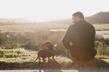 Man in black long sleeved shirt and blue jeans sitting out in nature next to a lake with a brown dachshund in a harness and on leash engaging with his pet dog. Sun flare. Lake and grass