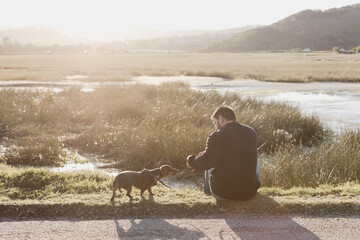 Man in black long sleeved shirt and blue jeans sitting out in nature next to a lake with a brown dachshund in a harness and on leash engaging with his pet dog. Sun flare. Lake and grass