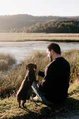 Man in black long sleeved shirt and blue jeans sitting out in nature next to a lake with a brown dachshund in a harness and on leash engaging with his pet dog. Sun flare. Lake and grass