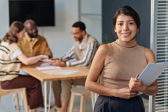 Portrait of young business leader with report smiling at camera standing at office with employees in background