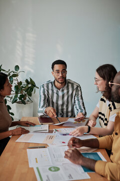 Young businessman presenting his report to other colleagues while they sitting at table and listening to him during meeting