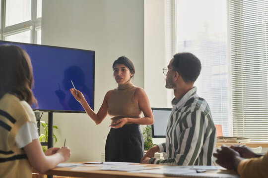 Young businesswoman pointing at big screen and presenting report to her colleagues during business presentation at office