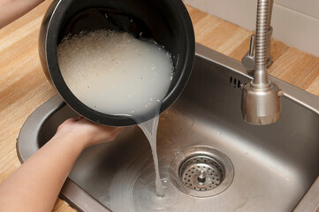 Person cleaning white rice in a rice cooker inner pot under running water, preparing it for...
