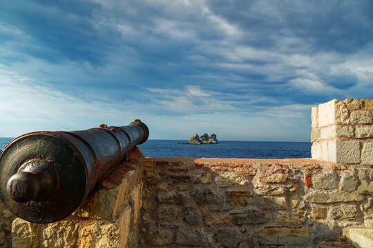 The old cannon on the walls of the Petrovac Fort, facing the sea, stands against the backdrop of a seascape with island covered by forest. Adriatic Sea, Montenegro