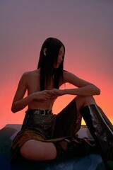 A young woman seated in a stylish outfit with a gradient background.