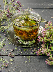 Homemade Oregano, Origanum vulgare herbal tea in a tea cup. Steaming hot drink with dry and fresh Oregano flower blossoms around, black color wood board minimal background.