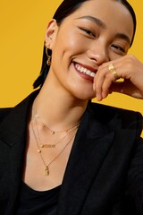 Smiling woman showcasing layered gold jewelry against a vibrant yellow background.