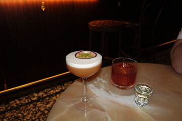 Stylish arrangement of three drinks — a passion fruit cocktail, a classic Negroni, and a clear shot — served on a polished marble table in a dimly lit upscale bar. Perfect for use in restaurant menus.