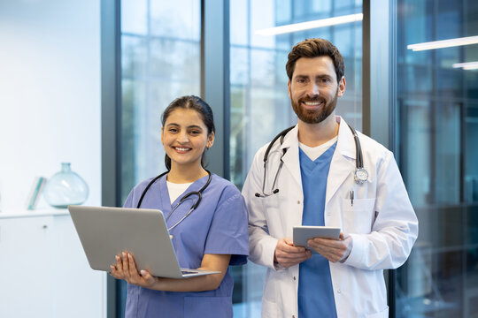 Two medical professionals, a doctor and a nurse, standing together using digital technology devices in a bright, professional office environment. They radiate teamwork and dedication