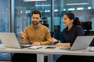 Two coworkers engage in a vibrant discussion while working on laptops in a bright, modern office...