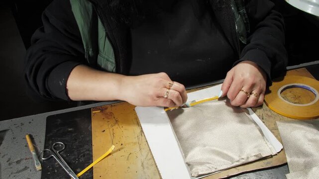 Woman tailor applies adhesive tape onto rolled up lining fabric workpiece at desk closeup. Seamstress makes casing inner cover in workshop