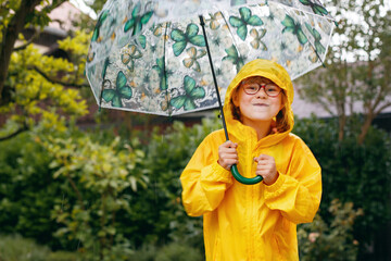 Little elementary school girl in yellow raincoat walking with big umbrella in rain. Happy child...