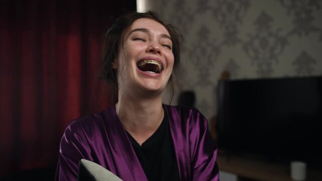 In a purple satin robe, a young woman with braces bursts into uncontrolled laughter, her eyes half-closed. The scene captures the fragile moment of an emotional breakdown, swinging between tears and