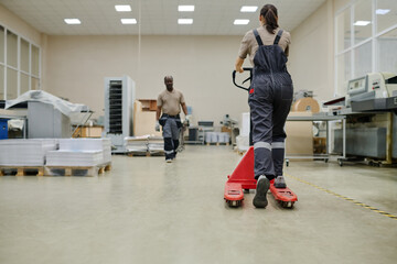 Horizontal rear view long shot of unrecognizable young woman wearing uniform using pallet jack at...