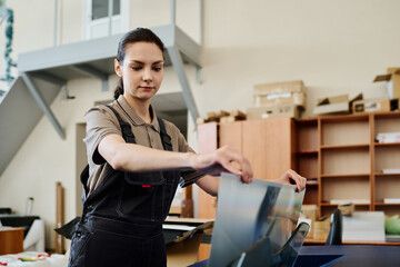 Medium portrait of young Caucasian female worker wearing apron printing poster in modern print shop