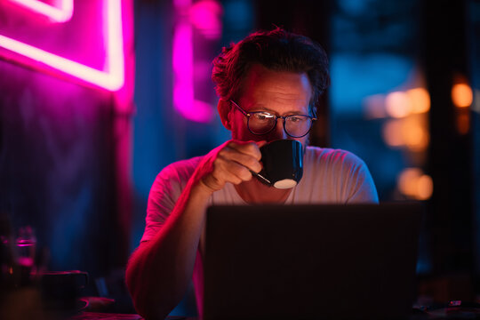 Man sips coffee while working late on his laptop, glowing in vibrant pink and blue neon light