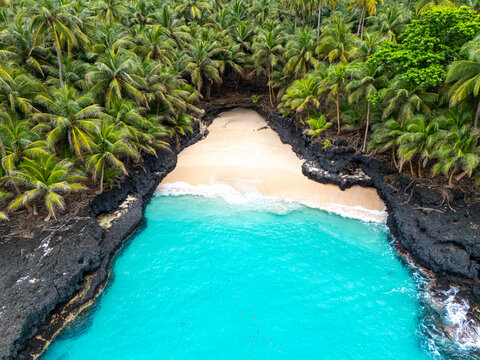 Tropical aerial view of Battery Beach on Rolas Island, S&atilde;o Tom&eacute; and Pr&iacute;ncipe, showcasing turquoise water, black volcanic rocks, and dense green palm forest.
