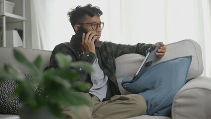 Asian man multitasking by talking on his phone while working on a tablet.