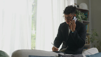 Asian man multitasking by talking on his phone while working on a tablet.