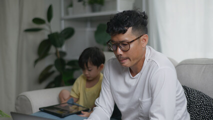 Asian father working from home on a laptop while his young son sits beside him using a tablet.