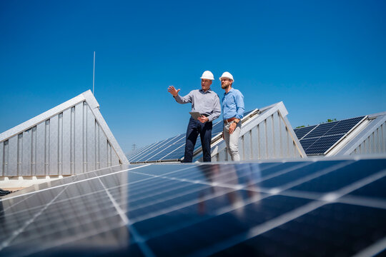Two businessmen wearing hardhats having a meeting on the roof of a building with solar panels