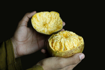 A close-up shot of a person's hands holding a halved ripe Bael fruit against a dark background,...