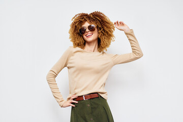 Smiling young woman with curly hair wearing sunglasses, posing confidently against a light gray...