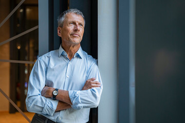 Thoughtful senior businessman standing with arms crossed in office corridor