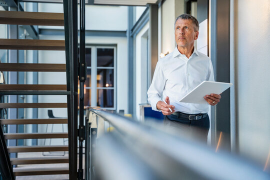 Thoughtful senior businessman standing with tablet PC in office corridor