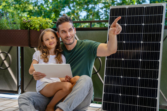 Father pointing with daughter holding tablet PC sitting next to solar panel in balcony