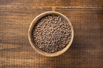 Wooden bowl filled with brown lentils placed on a wooden table, viewed from above.