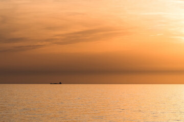 Cargo ship on the horizon during a golden sunset over the calm sea. Scenic seascape with warm orange sky and tranquil water.