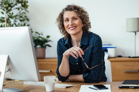 Smiling businesswoman holding eyeglasses and sitting at desk