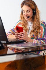 Young woman wearing a colorful sweater and headset working remotely at a desk with a laptop, checking her smartphone while multitasking, communication, and modern technology use.Vertical