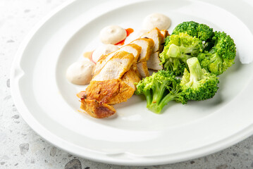 Grilled chicken breast with creamy sauce and steamed broccoli served on a white plate over a speckled countertop. Healthy homemade meal, top view.