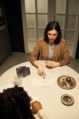 Caucasian young adult woman reading tarot cards for Black woman at round table, gesturing toward...