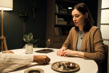 Caucasian young adult woman sitting at round table with Black woman, both engaging in tarot card...