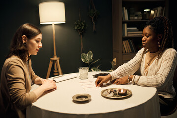 Caucasian young adult woman sitting at table with Black young adult woman reading tarot cards, both...