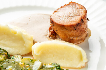 Gourmet pork tenderloin with creamy mashed potatoes, sliced cucumbers, herbs, and mustard seeds on a white plate with sauce, styled for elegant food presentation
