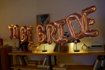 Metallic letter balloons spelling TO BE BRIDE hanging in decorated indoor setting, bookshelf with...