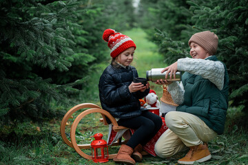 Cute little children decorating Christmas tree in the forest and drinking hot tea. Winter holidays,...
