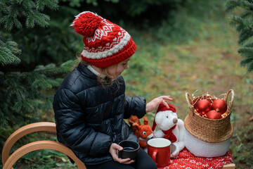 Cute little girl decorating Christmas tree in the forest and drinking hot tea. Winter holidays,...