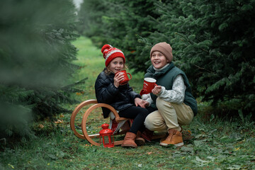 Cute little children decorating Christmas tree in the forest and drinking hot tea. Winter holidays,...
